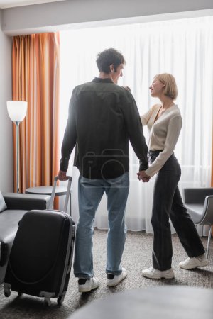 full length of man with travel bag holding hands with blonde smiling girlfriend in hotel room