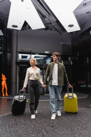 Foto de Full length of cheerful couple holding hands and looking away while walking with suitcases in lobby of modern hotel - Imagen libre de derechos