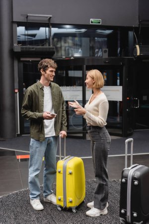 Foto de Young man with mobile phone smiling at blonde girlfriend talking near travel bags in hotel lobby - Imagen libre de derechos