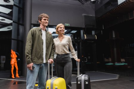 young man with smartphone and happy blonde woman looking away near travel bags in lobby of modern hotel