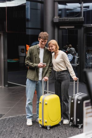 happy young couple with travel bags looking at mobile phone in hall of modern hotel