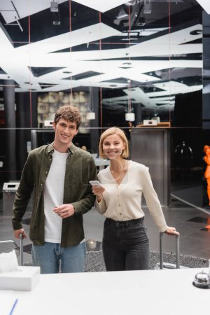 young man and cheerful blonde woman with mobile phone looking at camera near reception in modern hotel
