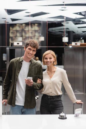 blonde woman and brunette man with smartphone smiling at camera near reception in modern hotel