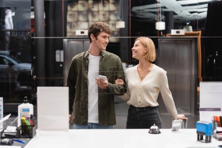 happy blonde woman and young man with cellphone looking at each other near reception in hotel