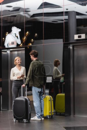 young blonde woman with smartphone smiling near boyfriend with suitcases in lobby of modern hotel