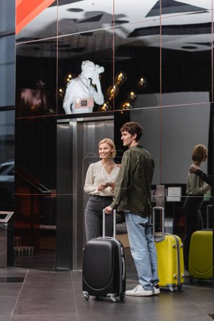 smiling couple with suitcases looking away while standing with travel bags in modern hotel