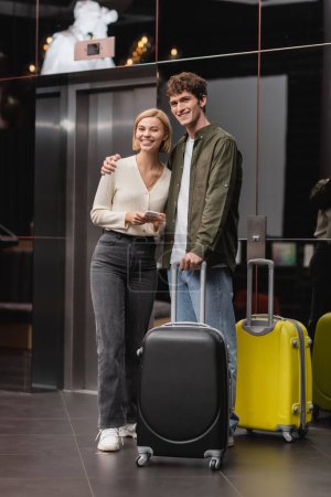 young man hugging joyful girlfriend with smartphone while standing near travel bags in hotel lobby