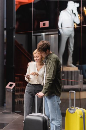 Téléchargez les photos : Joyful woman pointing at mobile phone near boyfriend and travel bags in hotel lobby - en image libre de droit