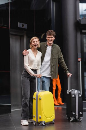 young man with blonde girlfriend standing with suitcases and laughing in lobby of hotel