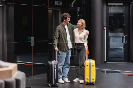 full length of young and happy couple smiling at each other near luggage in hotel lobby