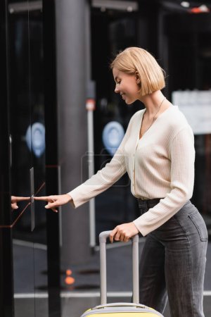 Photo pour Side view of smiling blonde woman with suitcase pressing elevator call button in modern hotel - image libre de droit