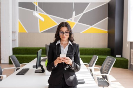 businesswoman in black suit and eyeglasses messaging on smartphone near desk with computers in modern office