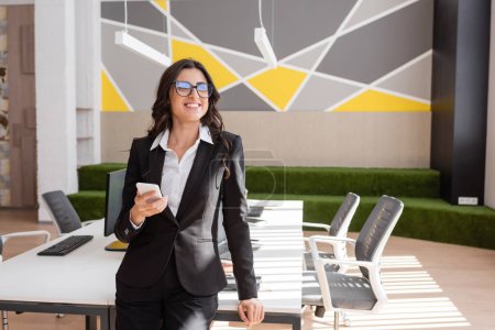 Foto de Happy businesswoman in black suit and eyeglasses holding smartphone and looking away in modern office - Imagen libre de derechos