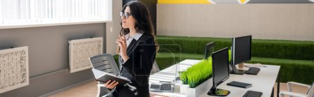 Téléchargez les photos : Brunette businesswoman in eyeglasses holding notebook while thinking near desk with monitors in office, banner - en image libre de droit