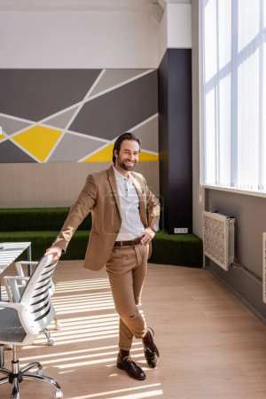 Téléchargez les photos : Full length of happy businessman in beige suit standing with hand on hip in spacious office - en image libre de droit