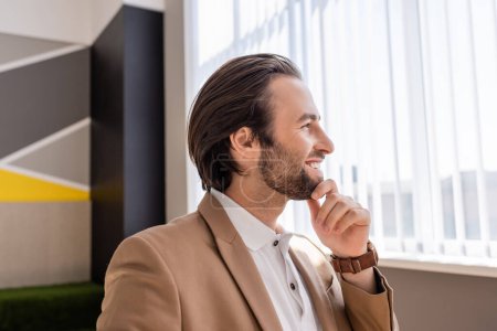 Foto de Smiling bearded businessman touching chin and looking away in office - Imagen libre de derechos