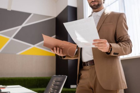 Téléchargez les photos : Partial view of bearded businessman in beige suit holding documents while working in office - en image libre de droit