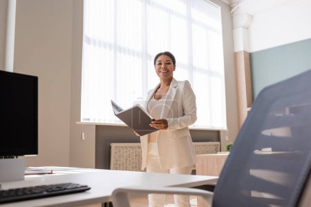 cheerful multiracial businesswoman with folder looking at camera near work desk in office