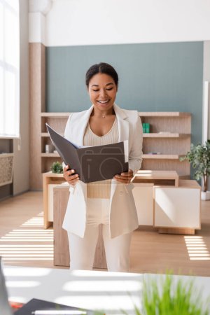 happy multiracial businesswoman looking at documents in folder while working in office