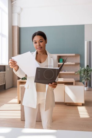 brunette multiracial woman in white blazer looking at documents in office