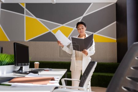 multiracial businesswoman working with documents while standing near desk in modern office