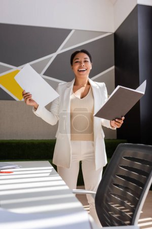 excited multiracial businesswoman in white suit holding documents and smiling at camera in office