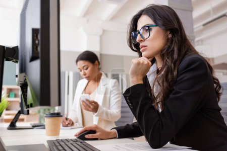 thoughtful businesswoman in eyeglasses working on computer near multiracial manager on blurred background