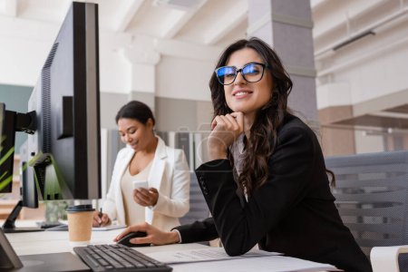businesswoman in eyeglasses smiling at camera near computer monitor and multiracial colleague on blurred background