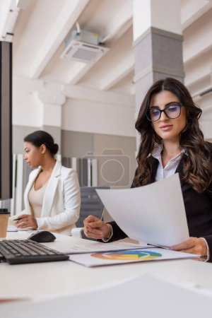 Téléchargez les photos : Businesswoman in eyeglasses working with documents near keyboard and multiracial colleague - en image libre de droit