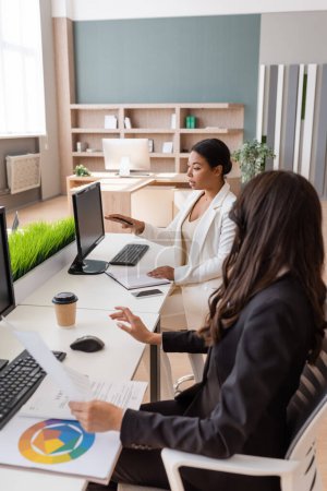 multiracial businesswoman pointing at computer monitor near colleague working with documents