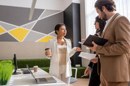 multiracial businesswoman with paper cup talking to colleagues near desk with computers in office