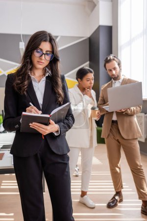 Photo for Businesswoman in eyeglasses writing in notebook near thoughtful interracial colleagues looking at laptop on blurred background - Royalty Free Image