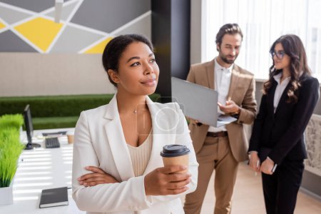 positive and thoughtful multiracial businesswoman standing with paper cup near colleagues with laptop on blurred background