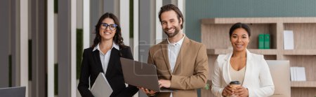 happy manager with laptop and interracial businesswomen with folder and coffee to go smiling at camera in office, banner