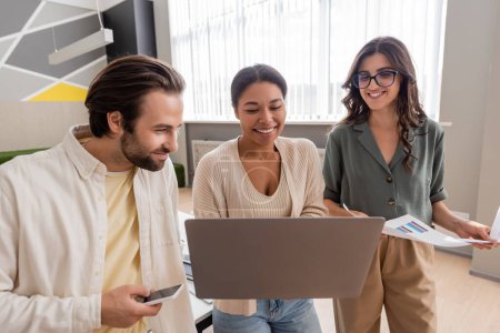multiracial businesswoman holding laptop near colleagues with infographics and smartphone in office