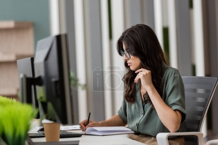 Photo pour Brunette manager lunettes écriture dans notebook près des moniteurs d'ordinateur sur flou au premier plan - image libre de droit