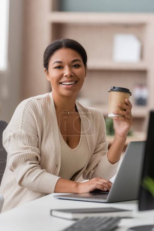 cheerful multiracial manager with takeaway drink looking at camera near blurred laptop in office