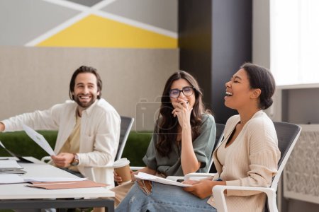 excited multiracial businesswoman sitting with documents and laughing with closed eyes near smiling colleagues in office
