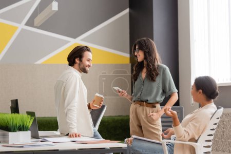 Foto de Young manager holding paper cup near smiling colleague with smartphone and multiracial businesswoman sitting in office - Imagen libre de derechos