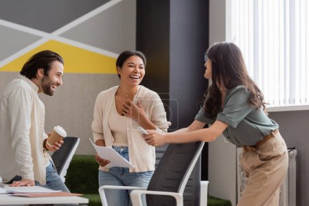 cheerful multiracial businesswoman holding papers and laughing near smiling managers in office