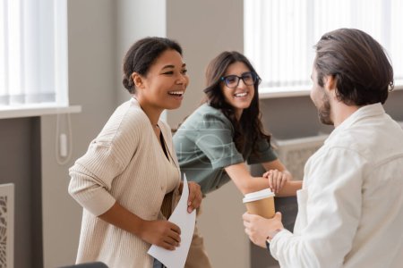 joyful interracial businesswomen laughing near young manager standing with coffee to go 