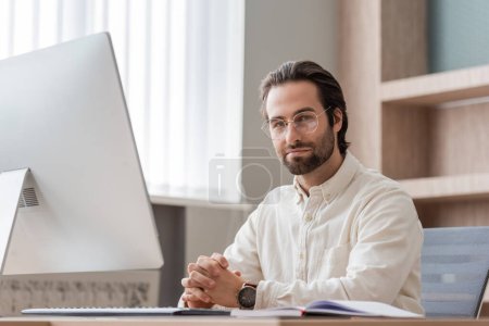 Foto de Young bearded businessman in eyeglasses sitting with clenched hands near computer monitor and looking at camera - Imagen libre de derechos