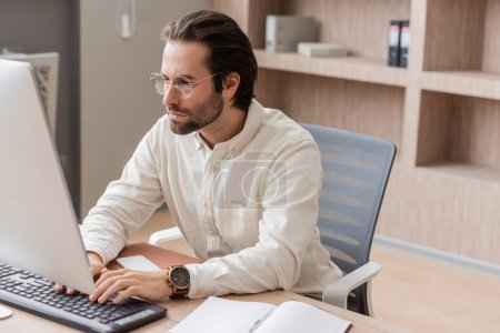 concentrated manager in eyeglasses working on computer near blank notebook on work desk