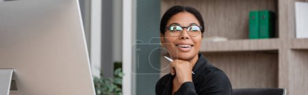 happy multiracial businesswoman in eyeglasses holding hand near chin and looking away in office, banner