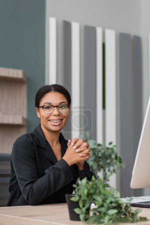 happy multiracial businesswoman in eyeglasses looking at camera near computer monitor and blurred flowerpot at workplace