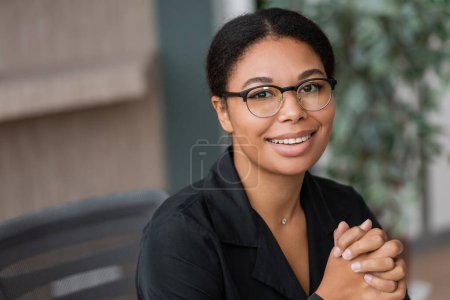 portrait of brunette multiracial businesswoman in eyeglasses smiling at camera in blurred office