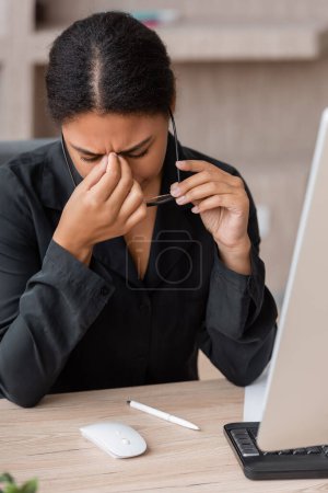 tired multiracial businesswoman holding eyeglasses and touching closed eyes while sitting at computer in office