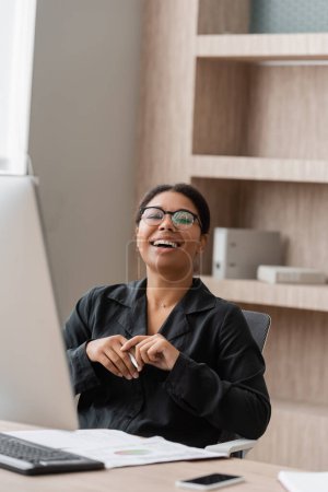 excited multiracial manager in eyeglasses looking at camera near computer monitor and documents in office