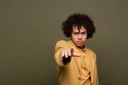 junger, lockiger Afroamerikaner in gelber Jacke, der mit dem Finger auf die Kamera zeigt, isoliert auf grau