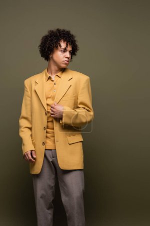 morena hombre afroamericano en blazer amarillo de moda y pantalones mirando hacia otro lado sobre fondo gris oliva